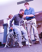 Dos empleados de un centro de vida asistida ayudan a un hombre mayor a levantarse de la silla de ruedas para que pueda caminar con andador