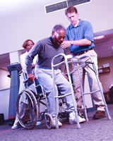 Two workers in an assisted living facility are helping an elderly man get out of his wheelchair so that he can walk with a walker