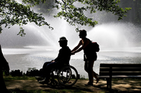 Una cuidadora lleva en silla de ruedas a su ser querido mientras pasean junto a un lago en un parque