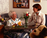 An elderly woman is in her room with flower arrangements and stuffed animals while talking to woman who is comforting her